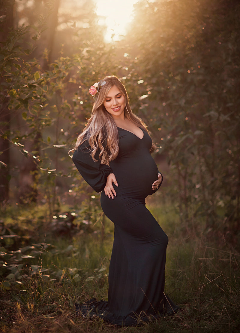 Pregnant woman wearing the Florence gown in blue spruce by Sew Trendy standing in grove of trees at sunset.