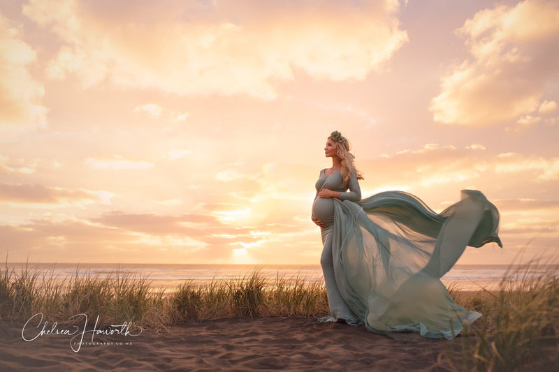Expecting mother wearing the Emerlie gown and detachable waterfall tossing train by Sew Trendy standing on the beach with her husband at sunset