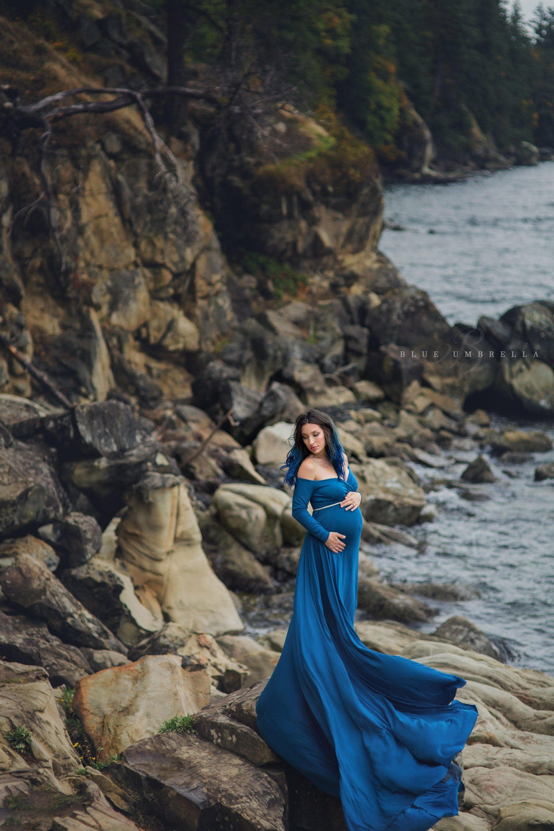 Pregnant woman wearing Bree gown in teal by Sew Trendy, standing on rocks next to ocean.