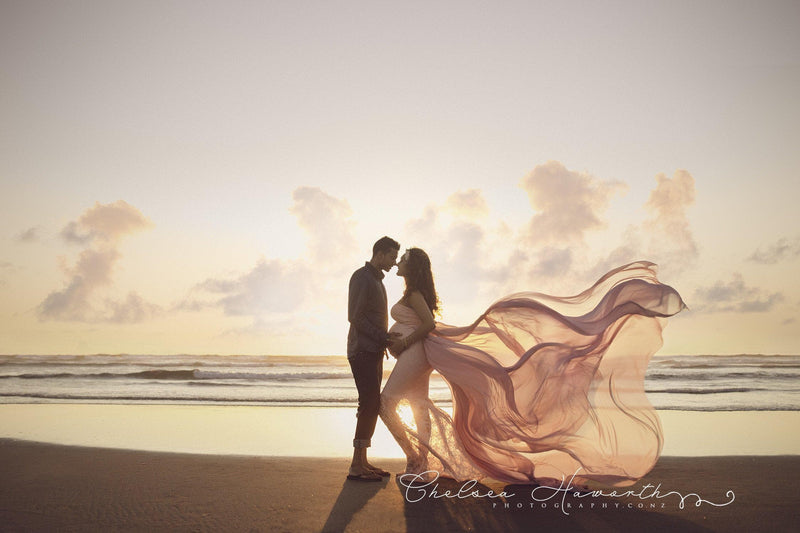 Expecting mother wearing the marilyn gown and detachable waterfall tossing train by Sew Trendy standing on the beach with her husband at sunset