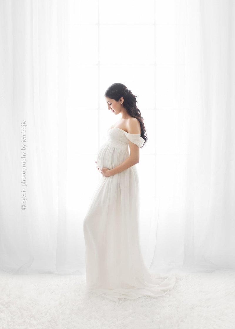 Pregnant mother wearing the Alina gown by Sew Trendy in ivory standing by a window 