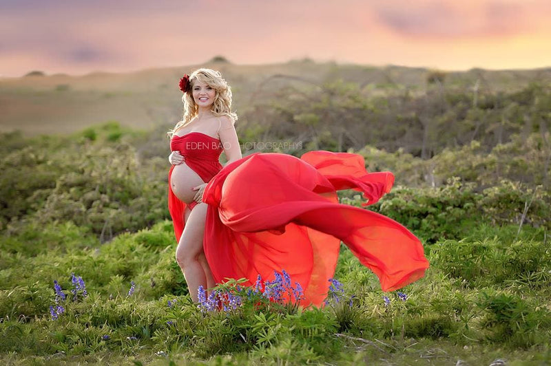 Pregnant woman in the Raquel Gown in Red by Sew Trendy Accessories standing in a field with flowers