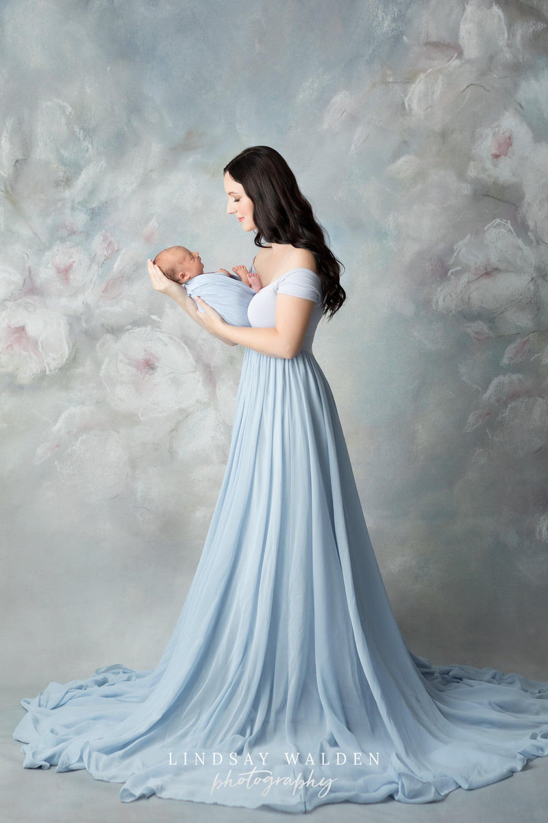 New mother holding her newborn and wearing the Faythe gown in blue rain by Sew Trendy standing in studio with floral backdrop