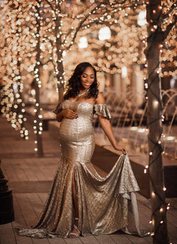 Pregnant woman in the Serenity Gown in Gold Sequin standing in front of a sidewalk with golden lights