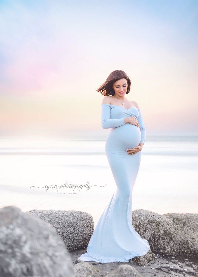 Pregnant woman wearing the Emerlie gown in blue rain by Sew Trendy standing on the beach