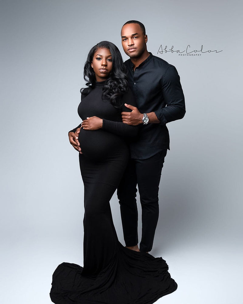 Photo of a beautiful straight black couple, dressed in all black on a light grey background. Woman is pregnant and wearing the Layne dress in black. The man is standing behind the woman and to the left. Her hands are resting on her belly and he is holding her on both sides.