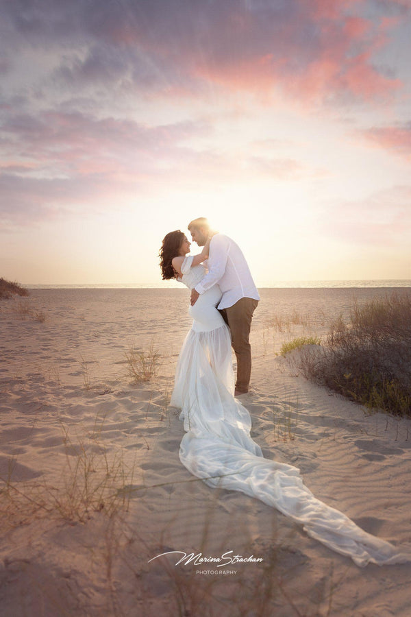 Expecting mother wearing the Angela gown in ivory by Sew Trendy, standing on beach embraced by husband.
