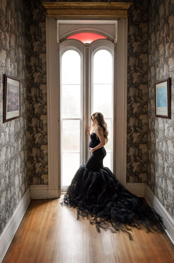 Pregnant woman wearing Celine gown in black by Sew Trendy standing near window in studio