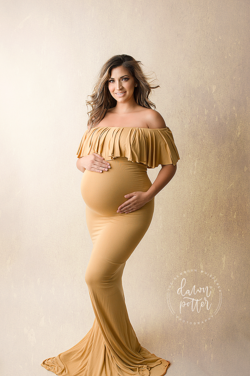 Expecting mother wearing the Colbie gown in gold by Sew Trendy standing in studio 