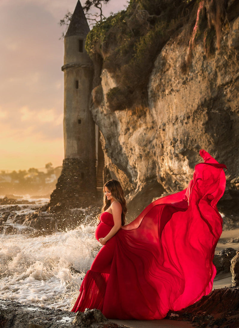 Pregnant mother in the Liv Gown by Sew Trendy Accessories in red standing on the beach with a castle in the background.