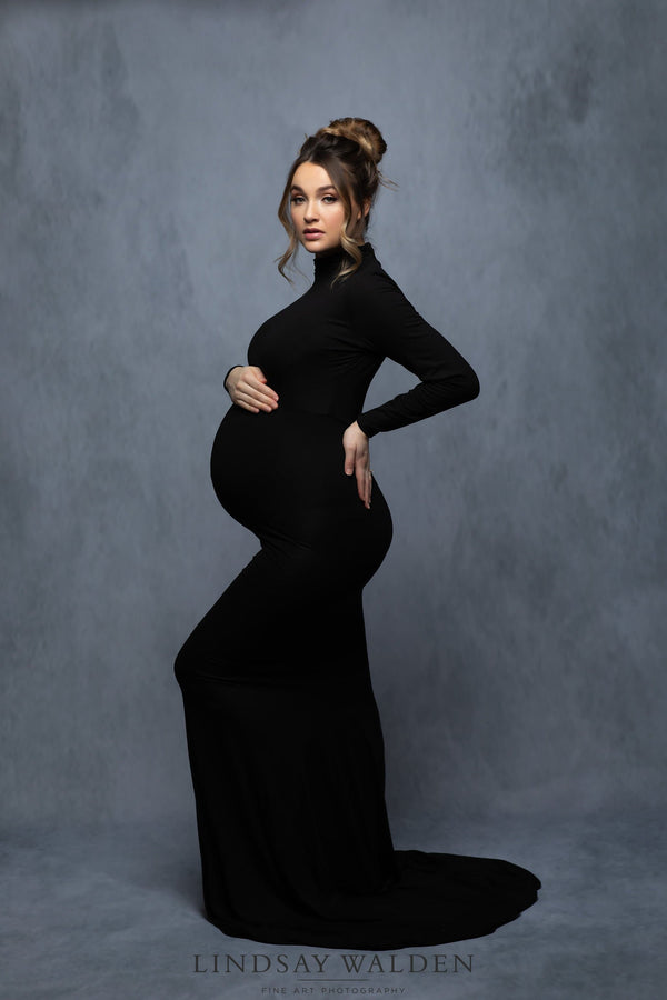 Gorgeous pregnant woman in a black and white photo wearing a black dress with long sleeves. She is resting her right hand on her belly and her left hand is on her hip. She is a very beautiful curvy female. She has long hair in an updo.