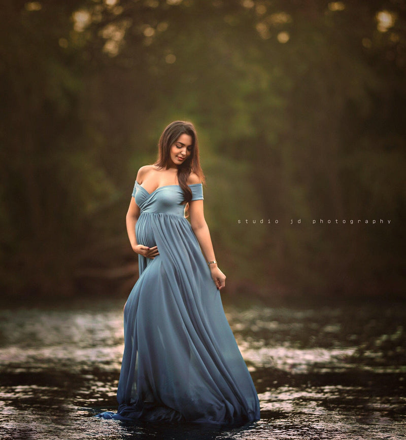 Expecting mother wearing the Faythe gown in steel blue by Sew Trendy standing in river at sunset