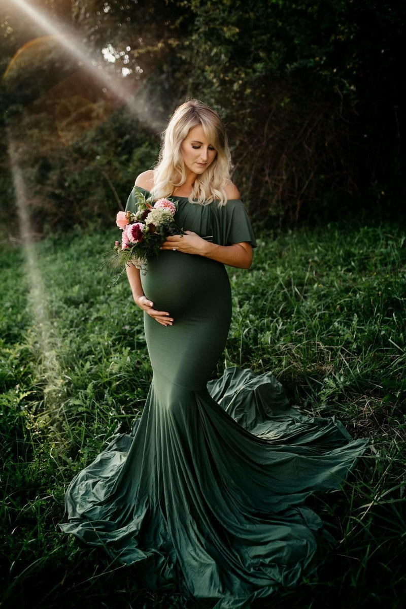 Pregnant woman wearing the Cirenya gown in evergreen by Sew Trendy, standing in field holding flowers.