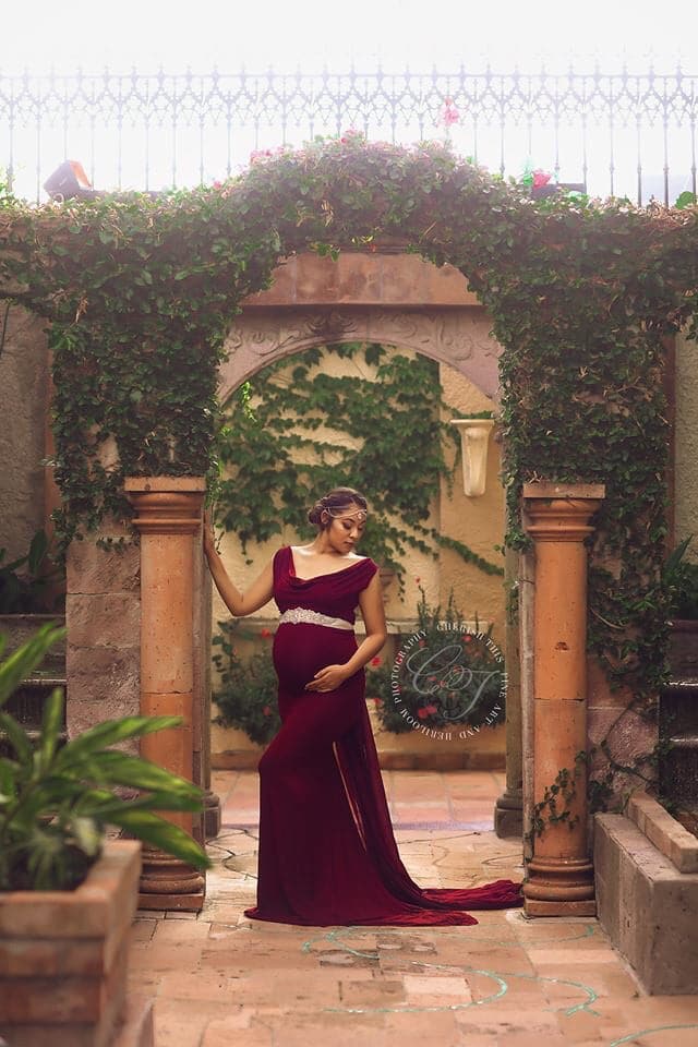 Pregnant woman in the Rue Gown in Burgundy by Sew Trendy Accessories standing in front of a stone archway.
