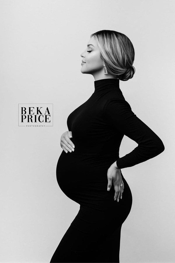 Gorgeous pregnant woman in black and white photo wearing a long black turtleneck gown with long sleeves. She is resting her right hand on her belly and left hand is on her hip. She is a very beautiful curvy female. She has highlighted hair in a bun.
