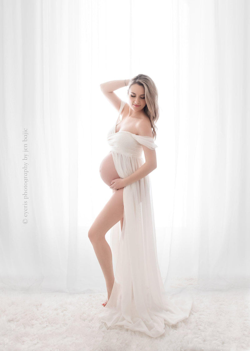 Pregnant mother wearing the Alina gown by Sew Trendy in ivory standing by a window 