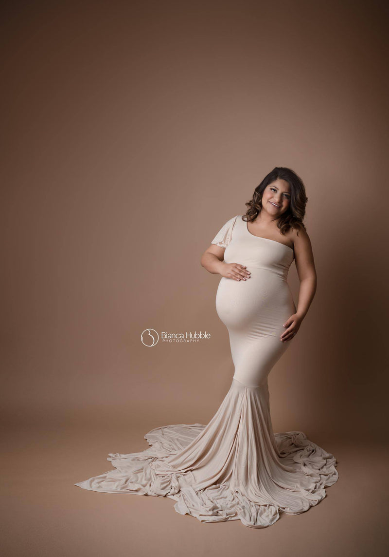 Pregnant woman wearing the Bonnie gown in champagne by Sew Trendy, standing in studio on pecan wallpaper.
