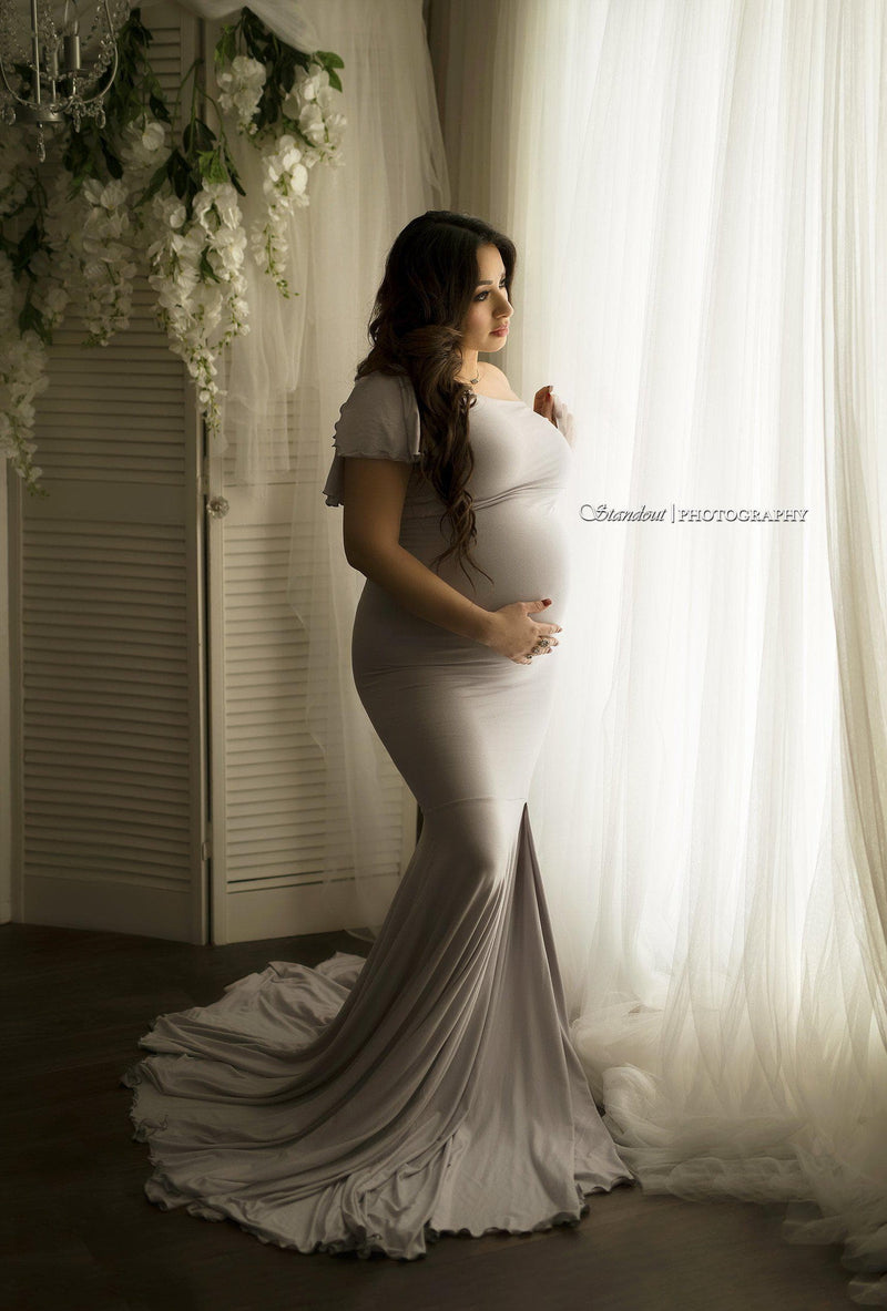 Pregnant woman wearing the Bonnie gown in grey by Sew Trendy, standing next to window with ivory curtains.