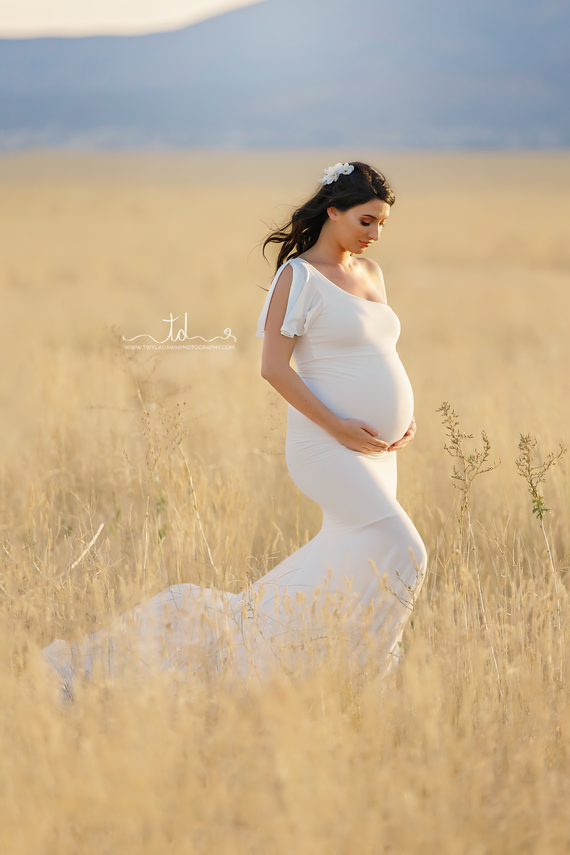 Pregnant woman wearing the Bonnie gown in ivory by Sew Trendy, standing in sunkissed wheat field.