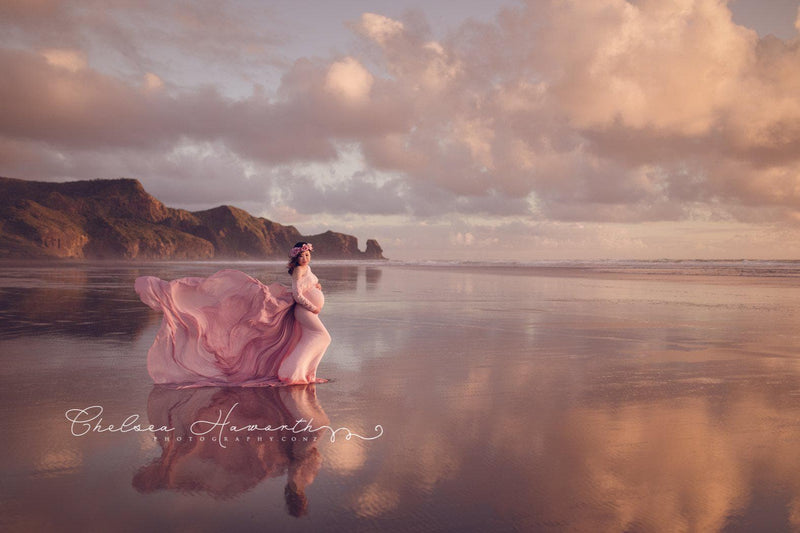Expecting mother wearing the Jaden gown and detachable waterfall tossing train by Sew Trendy standing on the beach at sunset