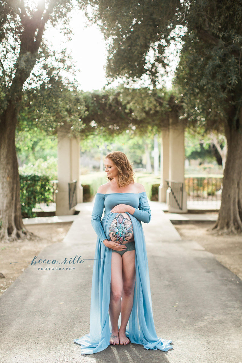 Pregnant woman in the Roxy Gown in Steel Blue by Sew Trendy Accessories Standing on a path in front of trees.
